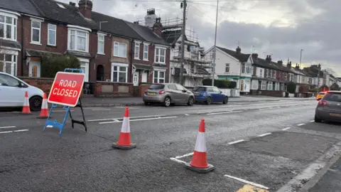 Sarah-May Buccieri/BBC Traffic cones and a "road closed" sign across residential street Carholme Lane. A police car with its lights on can be seen in the background.