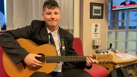 Matthew, a teenage boy, sit on a red sofa holding a light brown acoustic guitar. Smiling comfortably, he is wearing a black school uniform, including a black blazer, white shirt black tie with a red stripe.