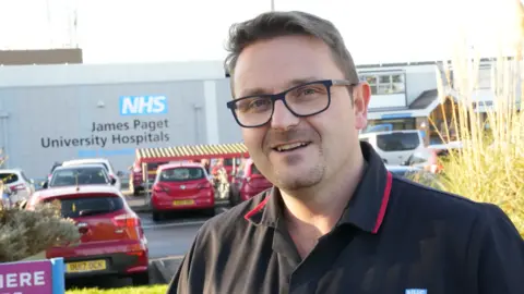 John Fairhall/BBC A man standing in front of a hospital. He is wearing glasses and a black polo-shirt. He is smiling at the camera.