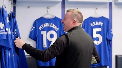 Everton FC Tony Sage is standing in the dressing room at Goodison Park. He is in front of Everton players' blue shirts, hanging up on coat hangers.