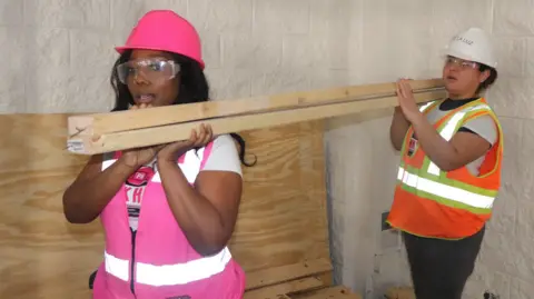 Traci Quinn Traci Quinn carrying a piece of wood with a worker