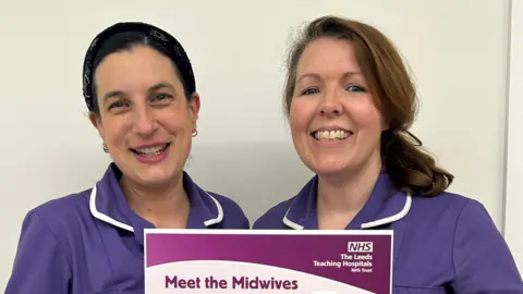 Two midwives in purple uniforms stand against a plain white wall, holding a poster about a new support session for expectant and new parents. 