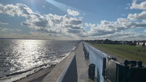 Image of the sea at Canvey Island. There is also a sea wall running down the centre into the distance. To the right of the wall is a grassy space. It is a bright sunny day with fluffy white clouds in the sky. A shard of light comes through the blue sky, from left to right. There are a small number of houses to the right of the grassy area. 