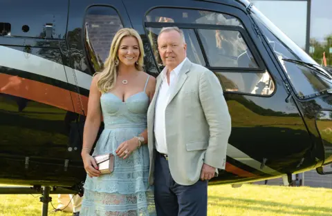 Ken Gaff Michelle Mone and Doug Barrowman in blue dress and casual grey jacket, jeans and white shirt respectively, stand in front of a brown helicopter.