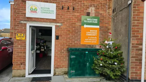 Jurate Matulioniene A medium-sized green Christmas tree is positioned on the right-hand side of a brick building. The building has a door on its right-hand side, which is open. The tree has been decorated with red and white baubles, silver stars and a miniature Christmas hat on top. 