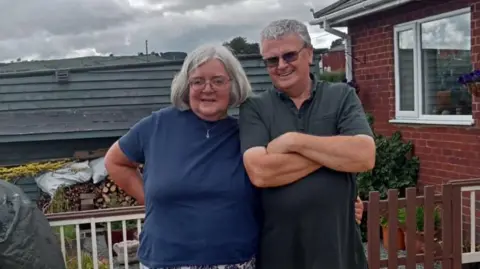 Family photo A woman with bobbed grey hair and a man with short grey hair are smiling while standing next to each other. They are on decking, with railing and a house behind them.