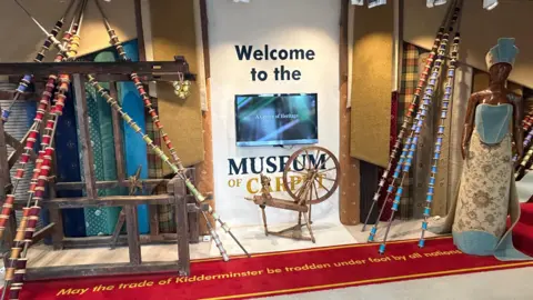 A display surrounding a flat screen TV. Words around the TV read "Welcome to the Museum of Carpets". Wooden contraptions are positioned around the area with material and displays also placed around the scene. 