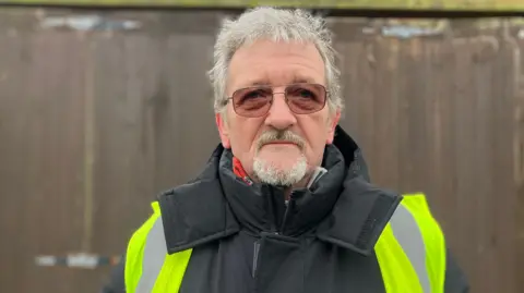 Martin Green, wearing warming clothing and a hi-vis vest, standing in front of a fence, looking at the camera