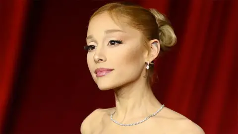 Getty Images Ariana Grande, a female singer and actress, at a gala. She is wearing a shiny necklace, looking into the distance with a red backdrop.