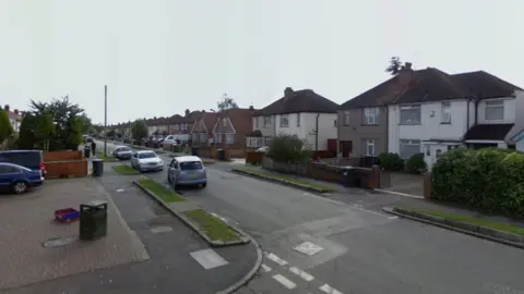 A screenshot from Google street view showing a residential street with semi-detached houses on one side and a short row of shops on the other. There are cars parked on one side of the street and it's a cloudy day.