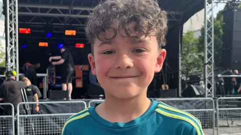 A young boy is stood outside smiling in front of a stage dimly lit behind him