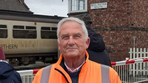 Glen Sanderson smiling into the camera. He has white hair and is wearing an orange hi-vis coat over a jumper and shirt. Behind him is the railway station which has a brick signalling tower on the right. A worn-looking grey Northern train is at the station. Two people stand behind him facing away from the camera in hats and coats. The railway barrier is down.