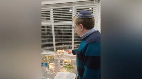 Rabbi Benjy Rickman Rabbi Benjy lighting the hanukkiah, he wears a striped jumper over a blue shirt and a blue yarmulke on his head.