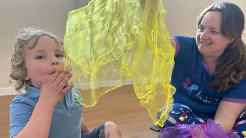 A little boy and his mum sit on wooden floor whilst playing with yellow and purple sheer scarves. The little boy has blonde hair and wears a light blue school polo top. He looks happy. The mother is wearing a dark blue and purple t-shirt with dark blue leggings. She has brown hair and is holding a yellow scarf above her sons head.