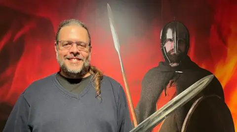 Discover Bucks Museum Brett Thorn standing in front of a life-size photograph of a man dressed as a Saxon warrior. Brett is on the left and has greying hair pulled back into a pony tail over his left shoulder. He is wearing glasses and holding up a replica sword, whose blade is pointing to the right.