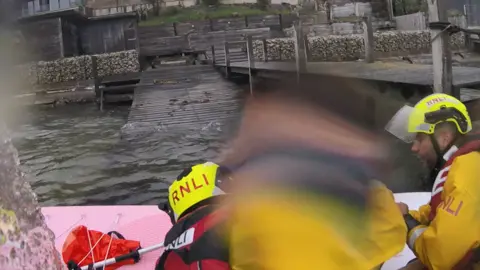 RNLI/Poole View from a lifeboat crew's camera showing colleagues in yellow overalls on board the lifeboat with a person in the centre who is blurred out.