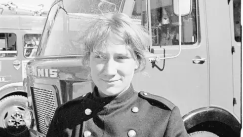 A young Mary Joy Langdon in a firefighter uniform. She is stood in front of a fire engine in the black and white image.