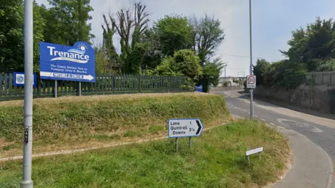 A blue sign with the word 'Trenance' on it in white writing. There is smaller text below it and an arrow pointing up to the entrance. There is a smaller white direction sign near the road on a grassy area. 
