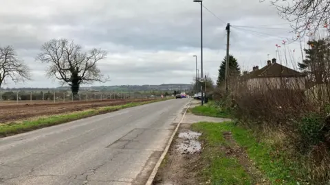 Radbourne Lane with fields surrounded by fencing on the left where houses are to be built and on the right are existing houses