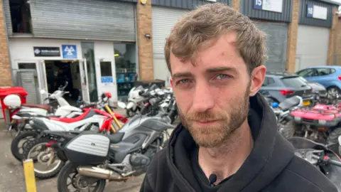 Jamie, who has short cropped brown hair and a beard. He is wearing a black hoodie and standing in front of a collection of motorbikes