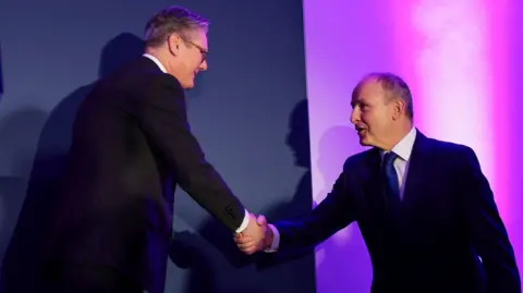 Micheál Martin said Sir Keir Starmer smiling and shaking hands in front of a blue backdrop with purple lighting. They are both wearing suits.