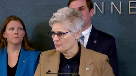 WCCO/CBS Mary Moriarty, waering a tan blazer, a black button up shirt and slightly tinted glasses, stands at a mircrophone, flanked by two other people