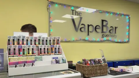 Shop interior, with large sign saying "The VapeBar Guernsey" on the wall behind the till. On the left of the till there is 3 rows of colourful vapes. On the right of the till there is a basket full of vapes.