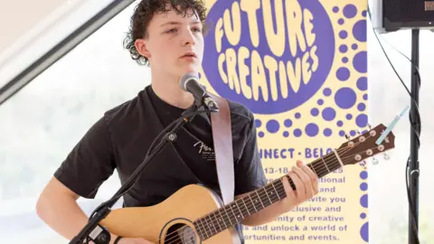 Callum O’Neil, who has wavy dark hair, is wearing dark top and holding a guitar. There is a microphone in front of him and a pull up banner behind him that reads 'Future Creatives'.