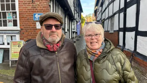 BBC An couple stand smiling on a narrow cobblestone street lined with historic black-and-white half-timbered buildings. He is in a flat cap, sunglasses and and brown leather jacket. She is wearing a gold-green coat and tartan scarf. 