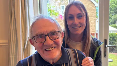 Kevin Shoesmith/BBC A bespectacled man with thinning grey hair is sitting down. A younger woman with long blonde hair gently rests a hand on his shoulder. Both of them are smiling.