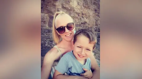 Laura Fricker Laura and Sebastian, looking about four years old, in the summer time by a brick wall on a beach. His mother is wearing her blonde hair back in a ponytail with large sunglasses and Sebby is being cuddled by his mum, is smiling cutely and wearing a blue T-shirt