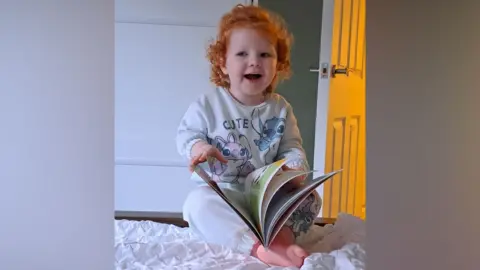 Joanne McShane Niamh reading a book. She has red curly hair, wearing grey Lilo and Stitch pyjamas.