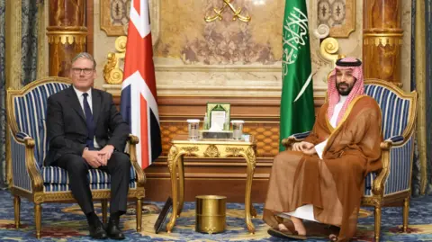 PA Media Prime Minister Sir Keir Starmer and Crown Prince Mohammed bin Salman are sitting on chairs with a gold-coloured table between them. They are sitting in front of the Union Jack and the flag of Saudi Arabia. 