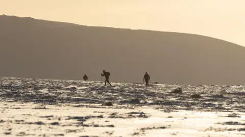 Jamie Rutherford / The Spine Race Three runners who are silhouetted in black travel in the ultramarathon Spine Race along the Pennine Way. It is either a morning or evening based off the sunlight. There is thick snow covering the landscape. 
