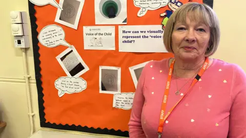 James Grant/BBC A woman in a pink jumper with hearts on, wearing a orange lanyard, stands in front of an orange board