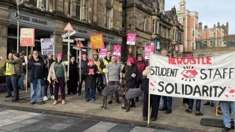 Dozens of picketers stand on the street holding up various placards. One says "Newcastle student staff solidarity", another reads "Strike against job cuts".