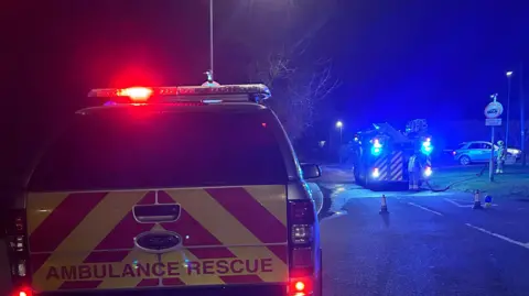 Kate Bradbrook/BBC An ambulance rescue vehicle in the foreground and, further away, a fire engine blocking off a road junction on a dark night. Two firefighters are at the back of the fire engine rolling up a hose.