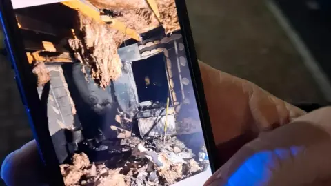 A hand holds a mobile photos showing photos of extreme fire damage to Chloe Taylor's home. 