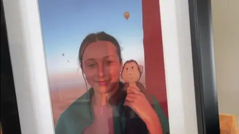 A framed photograph of Darcy smiling holding a small monkey teddy on her right shoulder. There are air balloons high in the sky behind her and the desert below.