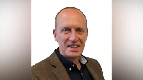 East Suffolk Council Paul Ashton is wearing a tweed jacket and black shirt and is smiling at the camera. The background is white.