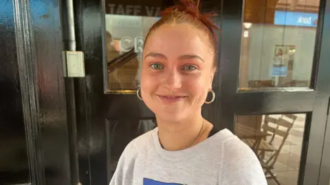 A young woman is smiling, looking at the camera. She has red hair tied up with hoop earrings and a lip piercing. She is standing outside a black door with glass panels.