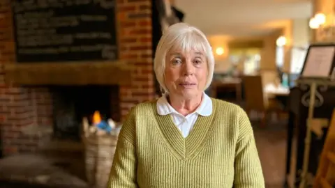 Martin Giles/BBC Maureen Jones is sitting in a pub. You can see a fireplace and a menu board on the wall. She is wearing a green-ish jumper and white shirt. 