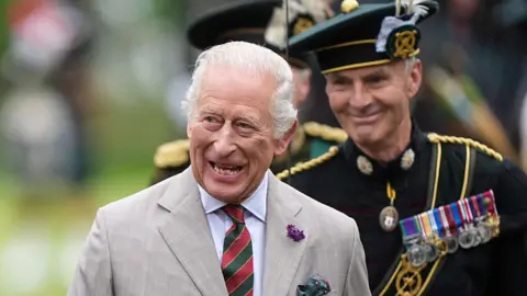 King Charles III wearing a grey suit with a white shirt and red and green striped tie. He is walking through the Palace Guard - a line of soldiers, wearing black uniforms.
