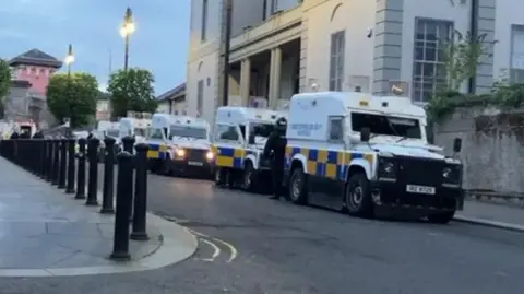 A number of police landrovers can be seen in a row near to Nailors Row in Londonderry. Black bollards line the other side of the road. A large pink building can be seen in the background as well.  