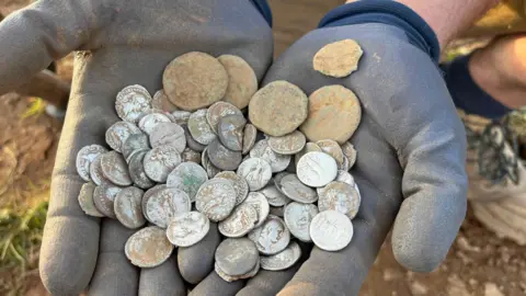 A pair of dirt-covered gloved hands hold a freshly unearthed hoard of worn silver and bronze Roman coins in an outdoor field setting.