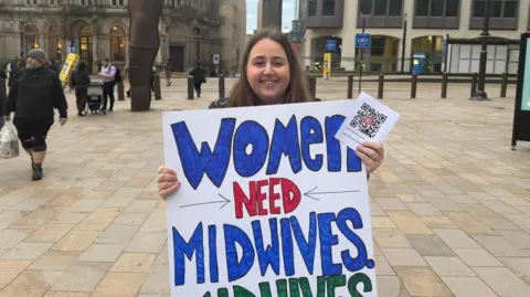 A woman with long dark hair and a smiling face holds a placard which reads "Women need midwives" the rest of the placard is not in view in this picture. She is standing on a square with pedestrians in the distance.