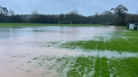 BBC A waterlogged, flooded pitch in Raglan, Monmouthshire. A white goal post can be seen at the other end of the pitch, with a row of trees beyond. 
