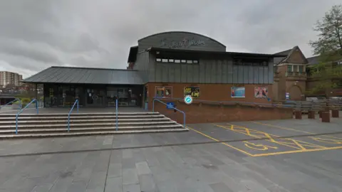 Google Exterior of the Electric Theatre in Guildford. Steps lead up to the main entrance and there is a sign saying 'Electric Theatre.' There are disabled parking spaces in front of the building.