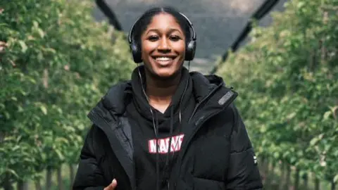 Thaliqua Smith Thaliqua Smith stands smiling at the camera, with hedges either side of her. She has her black hair tied up and is wearing a pair of black headphones. She has a black Marvel hoodie on, with a black puffer jacket over the top.