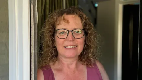 George King/BBC A woman with curly hair and glasses, wearing a pink vest top, smiling into the camera